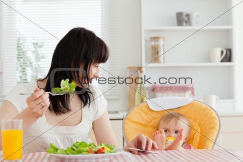 Beautiful brunette woman eating a salad next to her baby while s