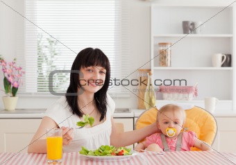 Charming brunette woman eating a salad next to her baby while si
