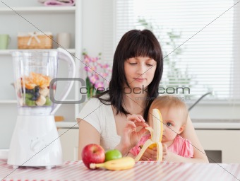 Cute brunette woman pealing a banana while holding her baby on h