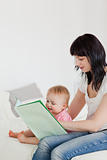 Cute brunette woman showing a book to her baby while sitting on 