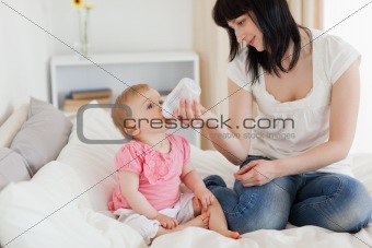 Charming brunette female bottle-feeding her baby on a bed