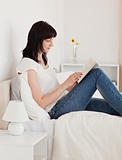 Pretty brunette woman reading a book while sitting on a bed