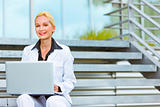 Smiling business woman sitting on stairs at office building and using laptop
