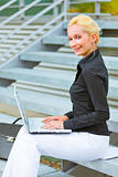 Smiling business woman sitting on stairs at office building and working on laptop

