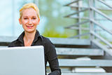 Pleased business woman sitting on stairs at office building and using laptop
