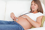 Charming pregnant woman using a stethoscope while lying on a sofa