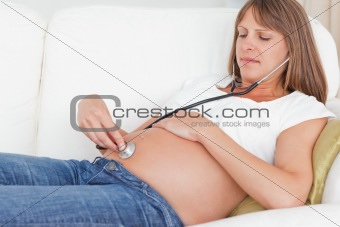 Charming pregnant woman using a stethoscope while lying on a sofa