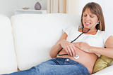 Attractive pregnant woman using a stethoscope while lying on a sofa
