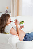 Beautiful pregnant woman eating a salad while lying on a sofa