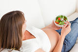 Charming pregnant woman eating a salad while lying on a sofa