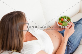 Charming pregnant woman eating a salad while lying on a sofa