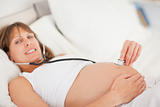 Pretty pregnant woman using a stethoscope while lying on a bed
