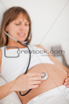 Attractive pregnant woman using a stethoscope while lying on a bed