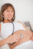 Lovely pregnant woman using a stethoscope while lying on a bed