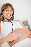 Nice pregnant woman using a stethoscope while lying on a bed