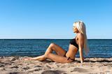 blonde girl in a black bathing suit sitting on shore blue sea 