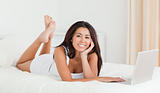 cute woman lying on bed with crossed legs and laptop looking int