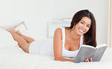 dark-haired woman lying on bed with book