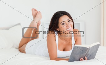 cute woman lying on bed with book looking into camera