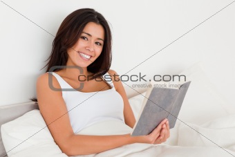 smiling woman holding book lying in bed looking into camera