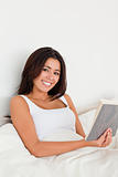cute woman holding book lying in bed looking into camera