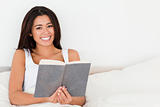 charming woman holding book lying in bed looking into camera