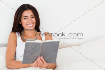 charming woman holding book lying in bed looking into camera