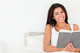 brunette woman holding book lying in bed looking into camera