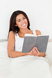 brunette woman holding book lyling in bed smiling into camera