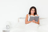 dark-haired woman holding book lyling in bed looking into camera