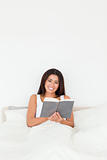 dark-haired woman holding book sitting in bed looking into camer
