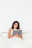 dark-haired woman reading book sitting in bed looking into camer