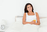 dark-haired woman holding orange juice sitting in bed looking at