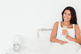 dark-haired woman dinking orange juice sitting in bed looking in