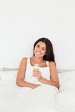 brunette woman holding orange juice sitting in bed looking into 