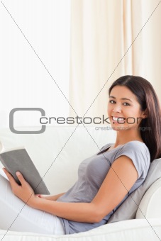 charming woman reading a book sitting on sofa smiles into camera