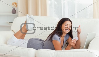 smiling woman with thumb up and card in hand lying on sofa looki