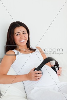 Frontal view of a pretty pregnant woman putting headphones on her belly while lying on a bed at home