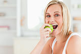 Young blonde woman sitting at table eating a green apple while l