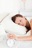 Sleepy red-haired woman lying in bed turning off alarm clock