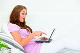 Confused pretty woman sitting on couch and pointing finger at laptop
