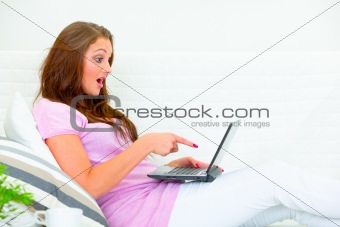 Confused pretty woman sitting on couch and pointing finger at laptop
