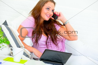 Happy pretty housewife on sofa talking on phone and using laptop
