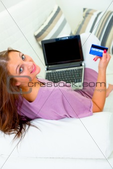 Smiling modern housewife sitting on sofa with laptop and credit card
