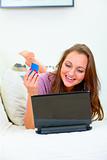 Cheerful beautiful woman lying on sofa and using credit card to shop from net
