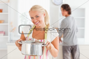 Woman posing with a boiler while her fiance is washing the dishes