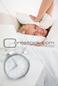Portrait of a tired woman hidding her head in a pillow while the alarmclock is ringing