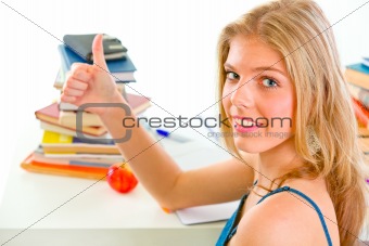 Smiling teen girl sitting at table with books and showing thumbs up gesture
