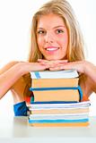 Smiling girl sitting at desk and holding hands on piles of books
