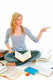 Cheerful teen girl sitting on floor among schoolbooks and pointing in corner
